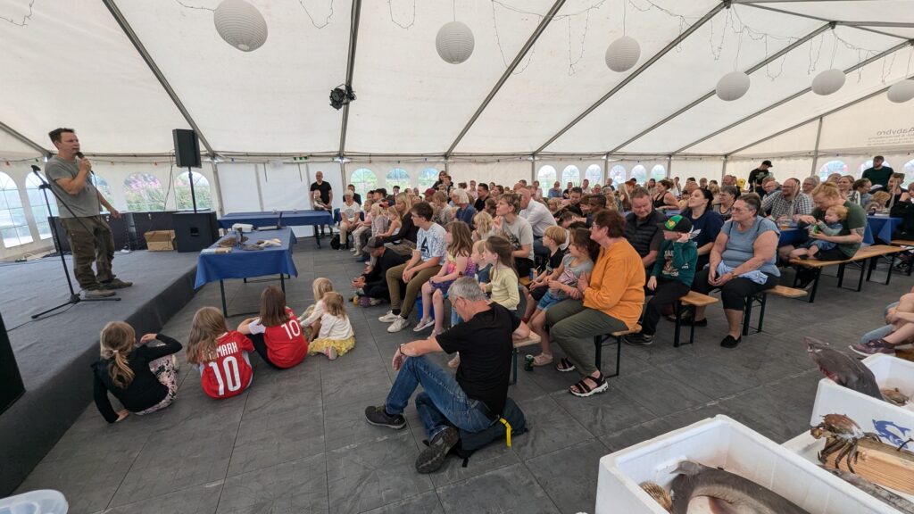 Anders Kofoed står på scenen og byder publikum velkommen.