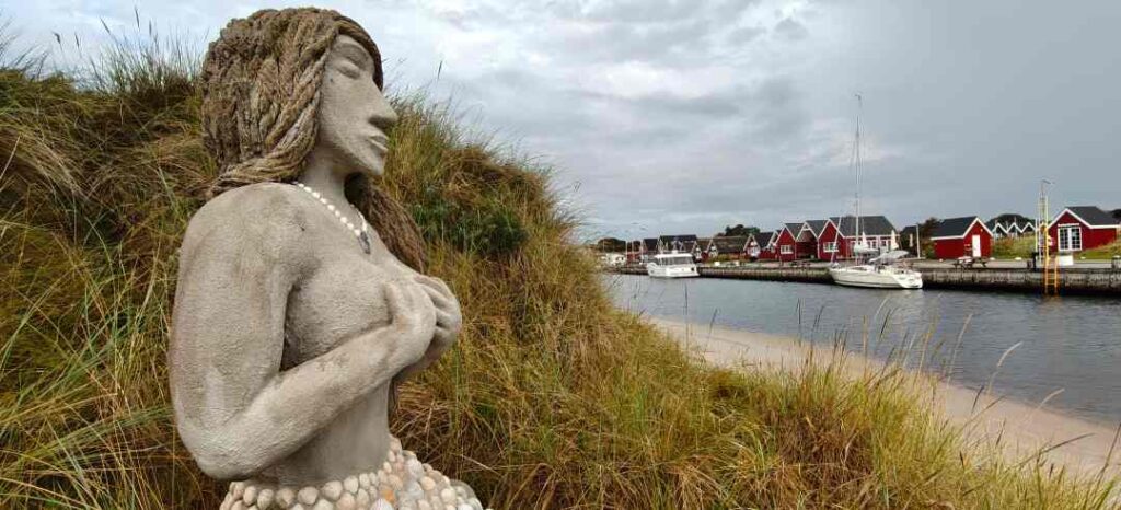 Havfruen Kaia sidder på en plads i klitterne syd for havnen i Hou og kigger ud mod havneindløbet.