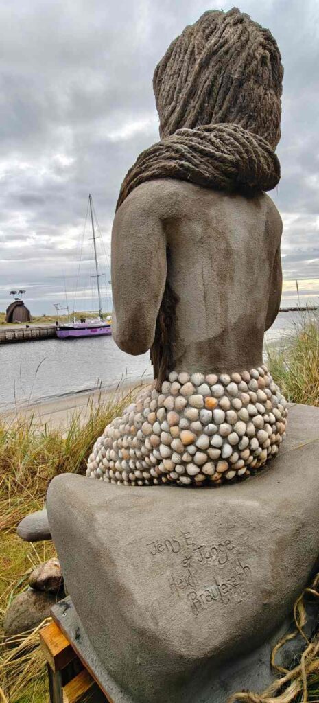 Havfruen Kaia sidder på en plads i klitterne syd for havnen i Hou og kigger ud mod havneindløbet. Her signeret af kunstnerne Jen Erik Junge og Heidi Brauteseth.