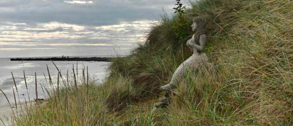 Havfruen Kaia sidder på en plads i klitterne syd for havnen i Hou og kigger ud mod havneindløbet.
